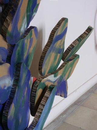 Nahaufnahme einer bemalten Pappskulptur, die Kaktusblättern ähnelt und vor einer weißen Wand und einem gefliesten Boden aufgestellt ist.