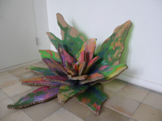 Eine große, farbenfrohe, pflanzenähnliche Skulptur aus geschichtetem, bemaltem Karton steht auf einem gefliesten Boden vor einer weißen Wand.
