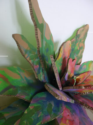 Eine große, mehrschichtige Pappskulptur, die abstrakten Pflanzenblättern ähnelt und in Grün-, Lila-, Rosa- und Orangetönen bemalt ist, steht vor einem schlichten weißen Hintergrund.