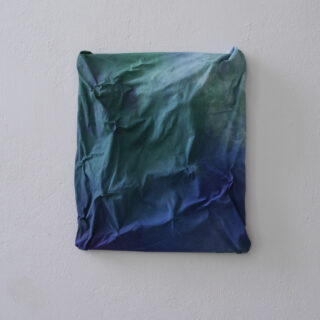 Ein zerknittertes, quadratisches Stück Stoff in Grün- und Blautönen ist an einer schlichten weißen Wand angebracht.
