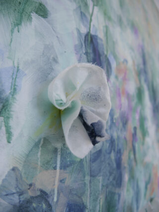 Eine Nahaufnahme einer weißen Stoffblume, die auf einem strukturierten, abstrakten, pastell- und blau bemalten Leinwandhintergrund befestigt ist.