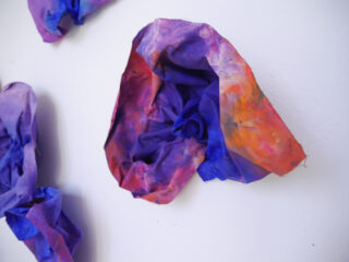 Ein in Lila-, Blau- und Orangetönen bemaltes Stück Stoff ist an eine weiße Wand gepinnt, wodurch eine strukturierte, dreidimensionale Form entsteht.