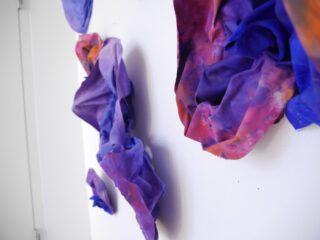 Stoffskulpturen in Lila-, Blau- und Rosatönen sind an einer weißen Wand befestigt und bilden eine strukturierte, dreidimensionale Kunstinstallation.