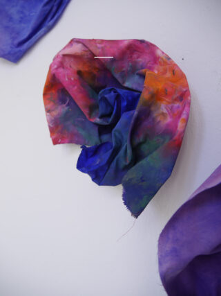 Ein Stück bunter, batikter Stoff ist locker gefaltet und an eine weiße Wand gepinnt. Es weist Blau-, Rot-, Rosa- und Grüntöne auf.