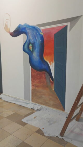 Ein surreales Wandgemälde zeigt eine blaue, abstrakte Form, die aus einer offenen Tür kommt und auf eine Wand gemalt ist. In der Nähe befinden sich auf dem gefliesten Boden eine Leiter und Abdeckplanen.