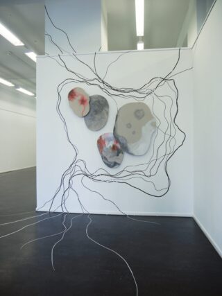 Drei abstrakte, steinartige Skulpturen mit roten und grauen Mustern sind an einer weißen Wand montiert, umgeben von schwarzen Linien, die sich auf den Boden und die angrenzende Wand erstrecken.