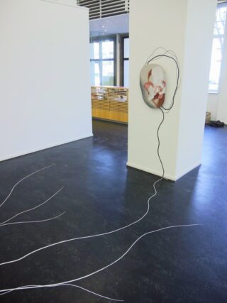 An einer weißen Wand ist eine Skulptur mit einer bemalten Gesichtsmaske angebracht; mehrere weiße Schnüre verlaufen von der Maske über den Boden und breiten sich in mehrere Richtungen aus.
