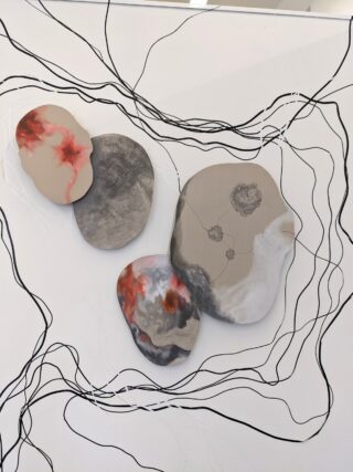 Vier abstrakte, unregelmäßig geformte Objekte mit grauen, schwarzen und roten Mustern sind an einer weißen Wand angebracht und durch schwarze Wirbellinien miteinander verbunden.