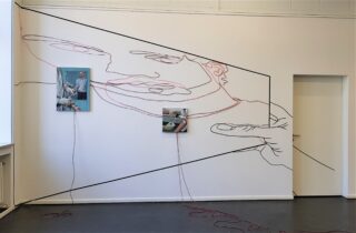 Eine Wandkunstinstallation zeigt eine große, durchgehende Strichzeichnung eines Gesichts und von Händen, dazwischen drei Fotos und eine rote Schnur, die vom Kunstwerk bis auf den Boden reicht.