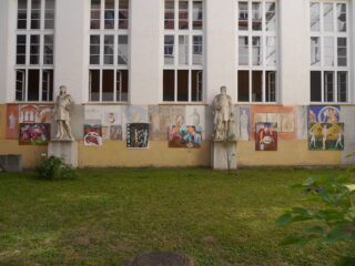 Ein Innenhof mit Gras, zwei Steinstatuen und farbenfrohen Wandgemälden an einer Wand unter großen Fenstern.