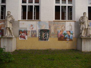 An einer gelben Wand unter zwei Steinstatuen und weiß eingefassten Fenstern an der Außenseite eines Gebäudes sind mehrere farbenfrohe Gemälde ausgestellt.