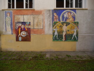 An der Außenwand eines Gebäudes sind unter zwei offenen Fenstern Gemälde und Wandmalereien mit religiösen und weltlichen Motiven zu sehen.