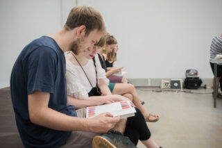 Mehrere Personen sitzen in einer Reihe im Innenbereich und betrachten Bücher oder Broschüren mit Farbmustern. Die Umgebung erinnert an eine Galerie oder ein modernes Klassenzimmer.