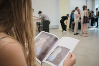 Eine Person liest eine Zeitschrift mit Fotos einer Person ohne Hemd, während sich andere Menschen in einem modernen Galerieraum versammeln und unterhalten.