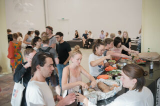 Eine Gruppe von Menschen versammelt sich bei einer gesellschaftlichen Veranstaltung in einem geräumigen, gut beleuchteten Raum um einen Tisch, an dem Essen serviert wird. Einige sitzen im Hintergrund, während andere sich unterhalten und essen.