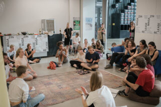 Eine Gruppe von Menschen sitzt im Innenbereich im Kreis auf dem Boden, einige klatschen, andere hören zu, im Hintergrund sind Plakate und Regale zu sehen.