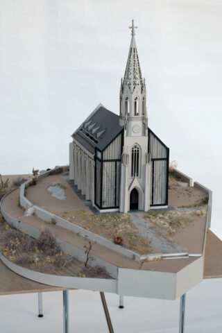 Ein detailliertes, maßstabsgetreues Architekturmodell einer Kirche im gotischen Stil mit einem hohen Turm, umgeben von Landschaftsgestaltung und Wegen auf einem Ausstellungstisch.