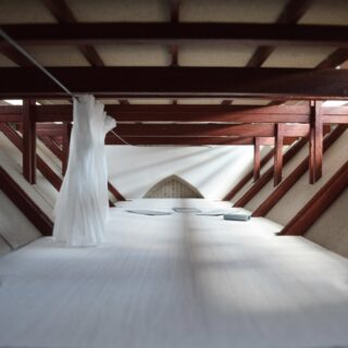 Ein geräumiger Dachboden mit freiliegenden Holzbalken, weißen Böden, einem hängenden Vorhang und verstreuten Papieren in der Nähe eines kleinen Bogenfensters am anderen Ende.