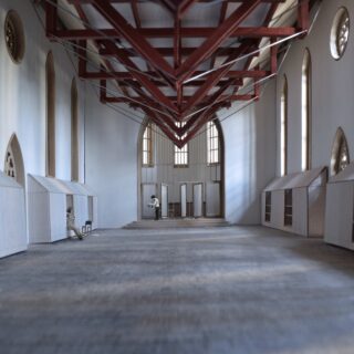 Eine geräumige Halle mit hohen Decken, freiliegenden roten Balken, hohen Fenstern und kleinen weißen Strukturen an beiden Seiten; am anderen Ende steht eine Person.