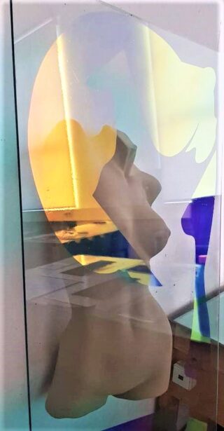 Eine reflektierende Glasplatte zeigt eine Büste im Seitenprofil und einen Teil eines Sonnenuntergangs am Meer, wobei die Merkmale der Skulptur mit farbenfrohen Reflexionen verschmelzen.