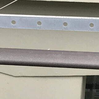 Eine Nahaufnahme eines Metallgeländers mit einer horizontalen Stange vor einer beigen Wand, über der sich ein perforierter Metallstreifen befindet.