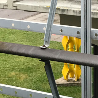 Ein Blick durch Metallgeländer und -balken, im Hintergrund sind eine Rasenfläche und eine gelbe abstrakte Außenskulptur zu sehen.