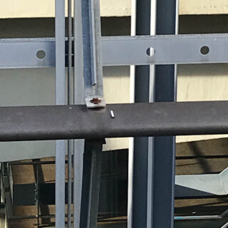 Nahaufnahme einer Metallstruktur mit Balken, von denen einer einen kleinen Rostfleck aufweist, und einem weißen zylindrischen Objekt, das auf einer horizontalen Oberfläche ruht.