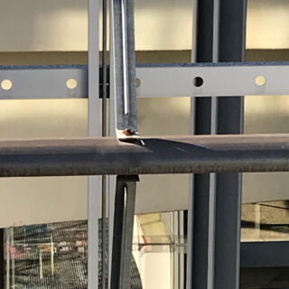 Nahaufnahme eines Metallträgers mit einer darauf ruhenden vertikalen Metallstange, positioniert vor einer Gebäudefassade mit Fenstern und Schatten werfendem Sonnenlicht.