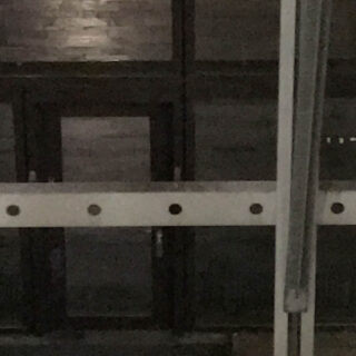 Dunkles, verschwommenes Bild von Metallträgern und einer reflektierenden Oberfläche, möglicherweise einem Fenster oder Boden, mit schwacher Beleuchtung und schattigen Bereichen.