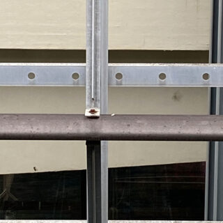 Eine Nahaufnahme eines Metallgeländers mit einem vertikalen Stützpfosten und horizontalen Balken vor einer cremefarbenen Wand und einem Glasfenster darunter.