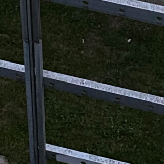 Nahaufnahme eines Metallzauns mit horizontalen Balken und Bolzen, der auf einer Rasenfläche steht.