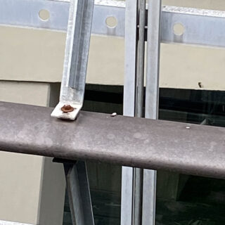 Eine Nahaufnahme eines Metallträgers, der an einer horizontalen Oberfläche festgeschraubt ist. Eine verrostete Schraube und Mutter sind sichtbar. Im Hintergrund sind ein beiges Gebäude und Glasplatten zu sehen.