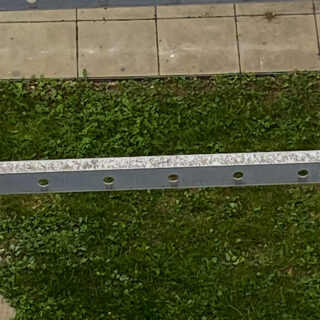 Ein Fliesenpflaster grenzt an ein Stück grünes Gras, über das ein horizontales Metallgeländer mit gleichmäßig verteilten Löchern verläuft.