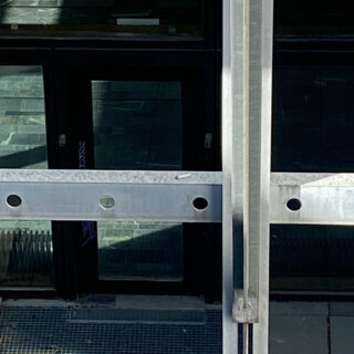 Vor dunklen Glastüren und einem gefliesten Boden befinden sich ein Metallgeländer und ein Stützbalken, wobei auf den Glasoberflächen schwache Reflexionen sichtbar sind.