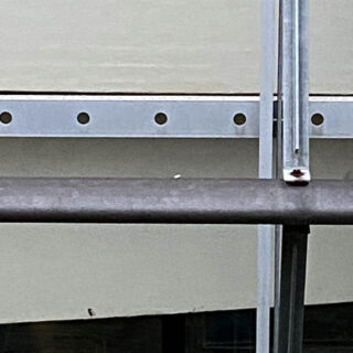Nahaufnahme eines Metallgeländers und Stützbalken vor einer cremefarbenen Wand mit gleichmäßig verteilten Löchern entlang eines horizontalen Metallstreifens.