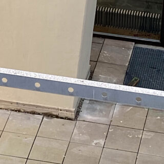 Ein gefliester Außenbereich mit einer beigen Wand, einer Glastür, einer Metallstange mit kreisförmigen Ausschnitten im Vordergrund und etwas verstreutem Bauschutt.