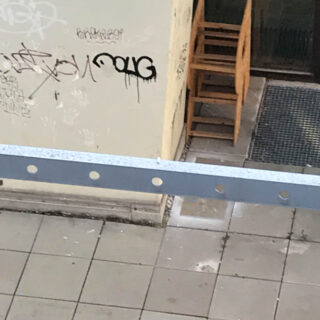Gestapelte Holzstühle neben einer Wand mit Graffiti, von oben gesehen; außerdem sind gefliester Bürgersteig und ein Metallgeländer zu sehen.