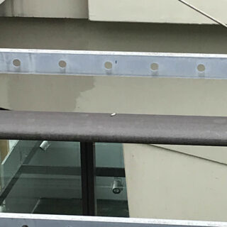 Eine Zigarettenkippe liegt auf einem braunen Geländer, im Hintergrund sind Metallträger und beigefarbene Gebäudewände zu sehen.