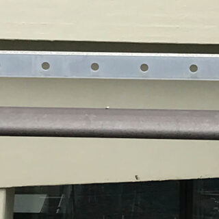 Eine Nahaufnahme einer Metallstange und eines perforierten Metallstreifens, die an einer beigen Wand angebracht sind. Unten ist ein Teil eines Fensters sichtbar.