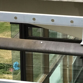 Eine einzelne weiße Pille liegt auf einem Metallgeländer vor einer Glasfassade eines Gebäudes mit Gras und einem blauen Objekt im Hintergrund.