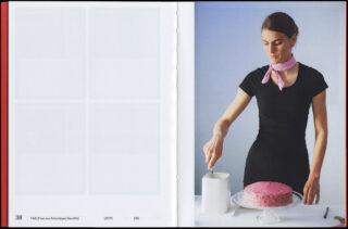 Eine Frau in einem schwarzen Kleid und einem rosa Schal verteilt mit einem Spatel rosa Zuckerguss auf einem runden Kuchen. Neben ihr stehen ein hoher weißer Behälter und eine Gabel auf dem Tisch.