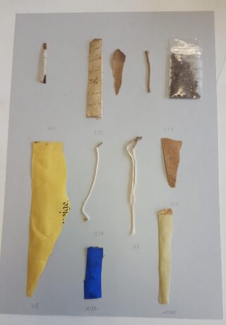 Eine Sammlung von Papier-, Schnur- und Stofffragmenten ist in Reihen auf einer flachen Oberfläche angeordnet, mit handschriftlichen Nummern neben jedem Gegenstand.