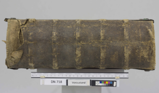 Ein großes, abgenutztes, in Leder gebundenes Buch liegt mit dem Rücken nach oben und sichtbaren Rissen da; darunter ein Etikett mit der Aufschrift „DN 718“ sowie einer Farbskala und einem Lineal als Referenz.