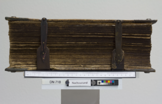 Ein großes, altes Buch mit verwitterten Seiten und zwei Metallschließen, aufrecht auf einem Ständer positioniert, mit Lineal, Farbskala und der Aufschrift „DN 718 Nachzustand“.