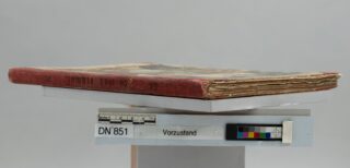 Ein abgenutztes, dünnes Hardcover-Buch mit rotem Rücken und ausgefransten Seiten liegt auf einer weißen Unterlage neben einem Lineal und einer Farbkalibrierungstabelle mit der Aufschrift „DN‘ 851 Vorzustand“.