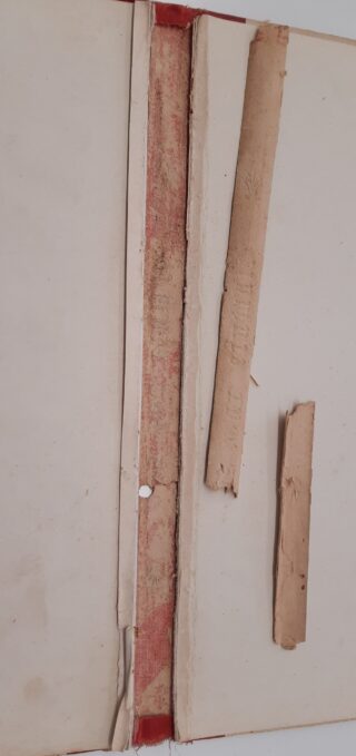 Ein Buch mit freiliegendem und abgelöstem Rücken; zwei abgebrochene Stücke des Rückens liegen auf der rechten Seite des offenen Einbands.