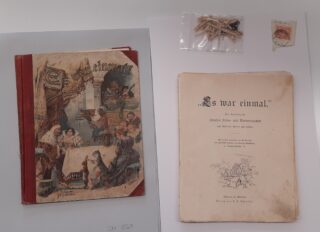 Auf einer weißen Fläche sind ein altmodischer Bilderbuchband, ein passendes Titelblatt, ein kleines Säckchen mit zerbrochenen Streichhölzern und eine Briefmarke angeordnet.