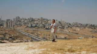Eine Person steht auf einem trockenen, grasbewachsenen Hügel und überblickt unter klarem Himmel eine geteilte Stadtlandschaft mit Straßen, einer Sperrmauer und dicht bebauter Bebauung im Hintergrund.