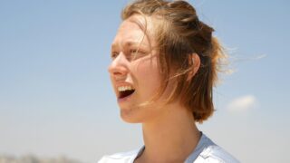 Eine Person mit kurzen Haaren steht draußen im Sonnenlicht, den Mund geöffnet, als würde sie sprechen oder singen, mit einem blauen Himmel im Hintergrund.
