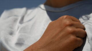 Eine Nahaufnahme der zur Faust geballten Hand einer Person, die diese an ihre Brust hält. Vor einem blauen Hintergrund sind Teile eines weißen Hemdes und des Halses zu sehen.